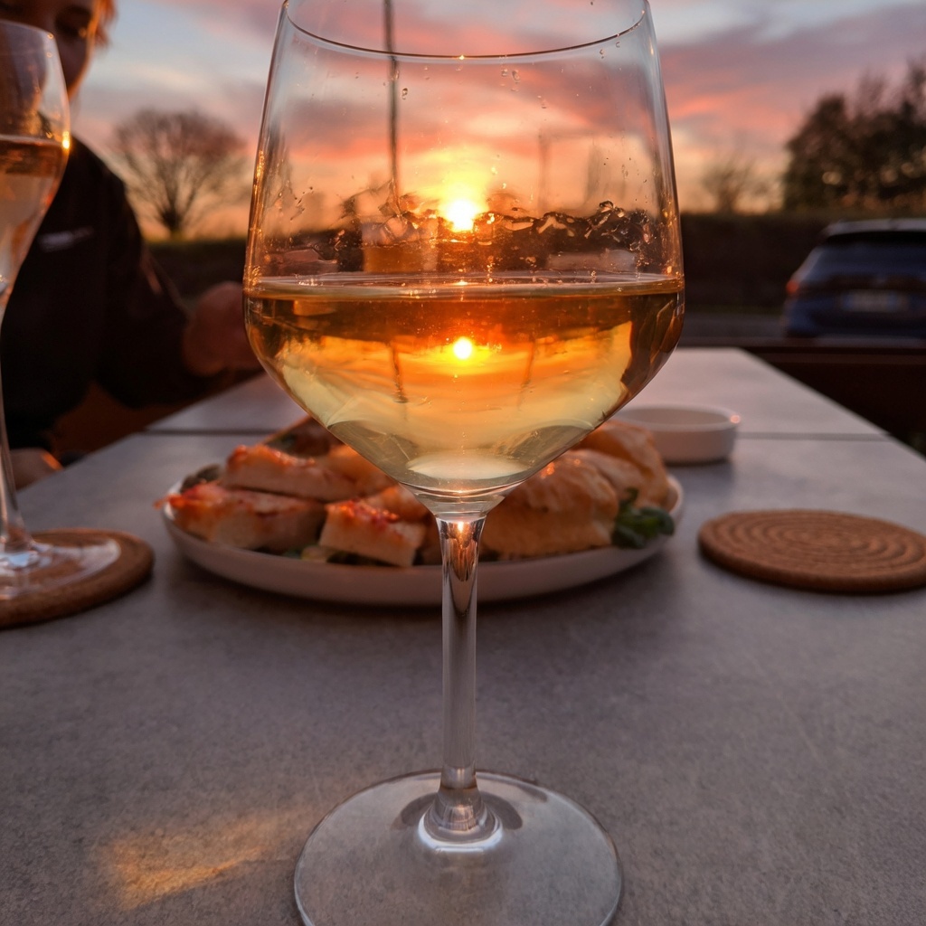 Calice di vino bianco al tramonto su tavolo all'aperto, con piatto di focaccia al pomodoro e sottobicchieri in fibra
