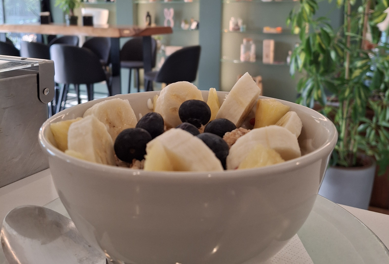 Ciotola bianca con banana, ananas, mirtilli e granola su un tavolo, sedie nere e scaffali verde salvia sullo sfondo